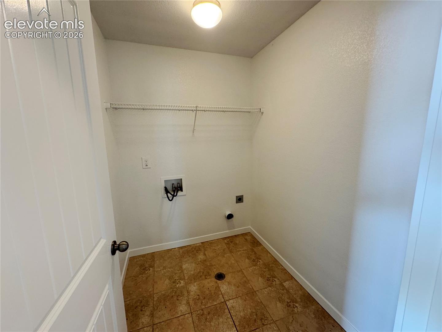 Utility area featuring tile flooring, ceiling-mounted lighting, wall-mounted wire shelving, and dedicated water and electrical hookups