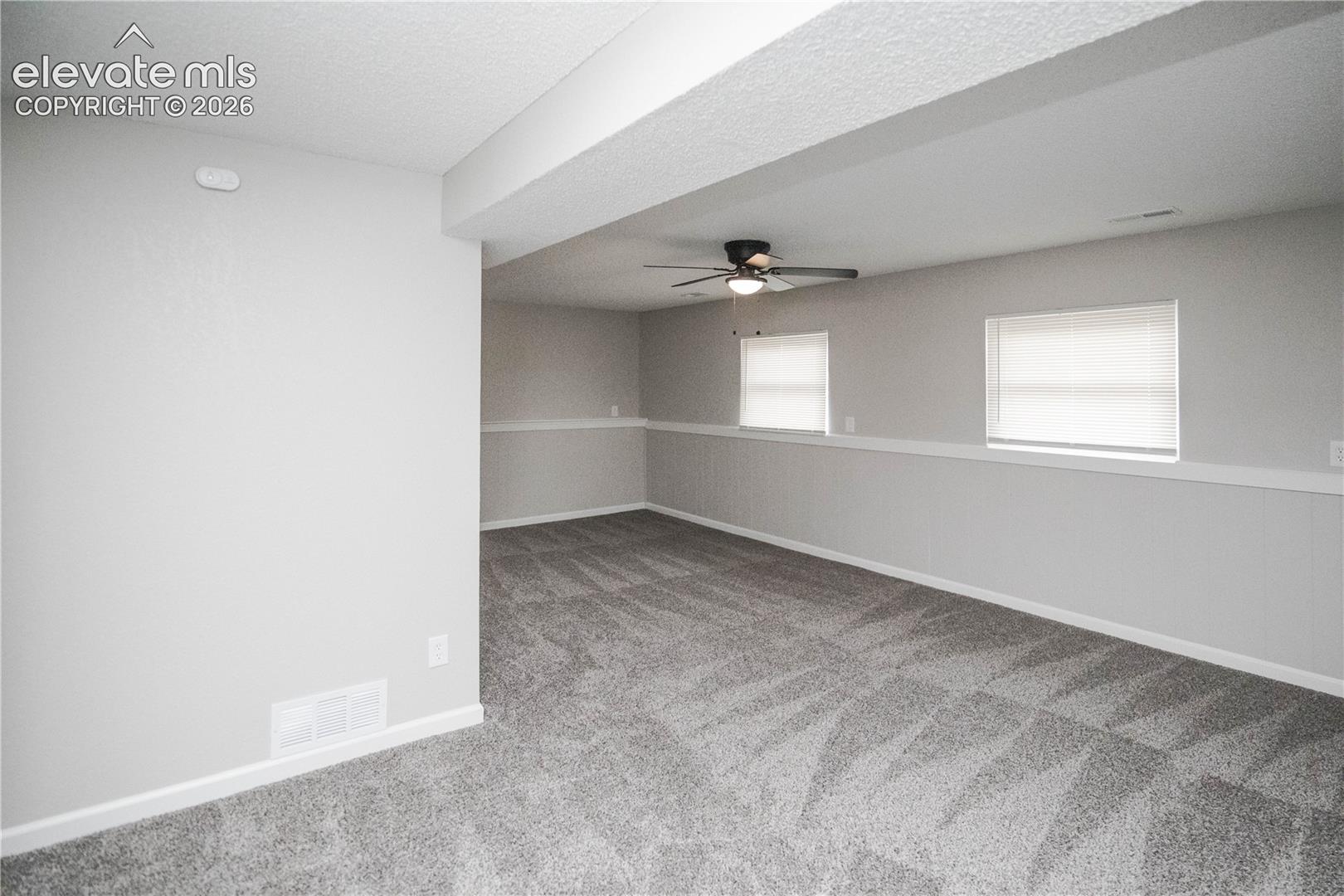 Neutral-toned interior space featuring wall-to-wall carpeting, white chair rail molding, dual windows with integrated blinds, and a central ceiling fan with integrated lighting