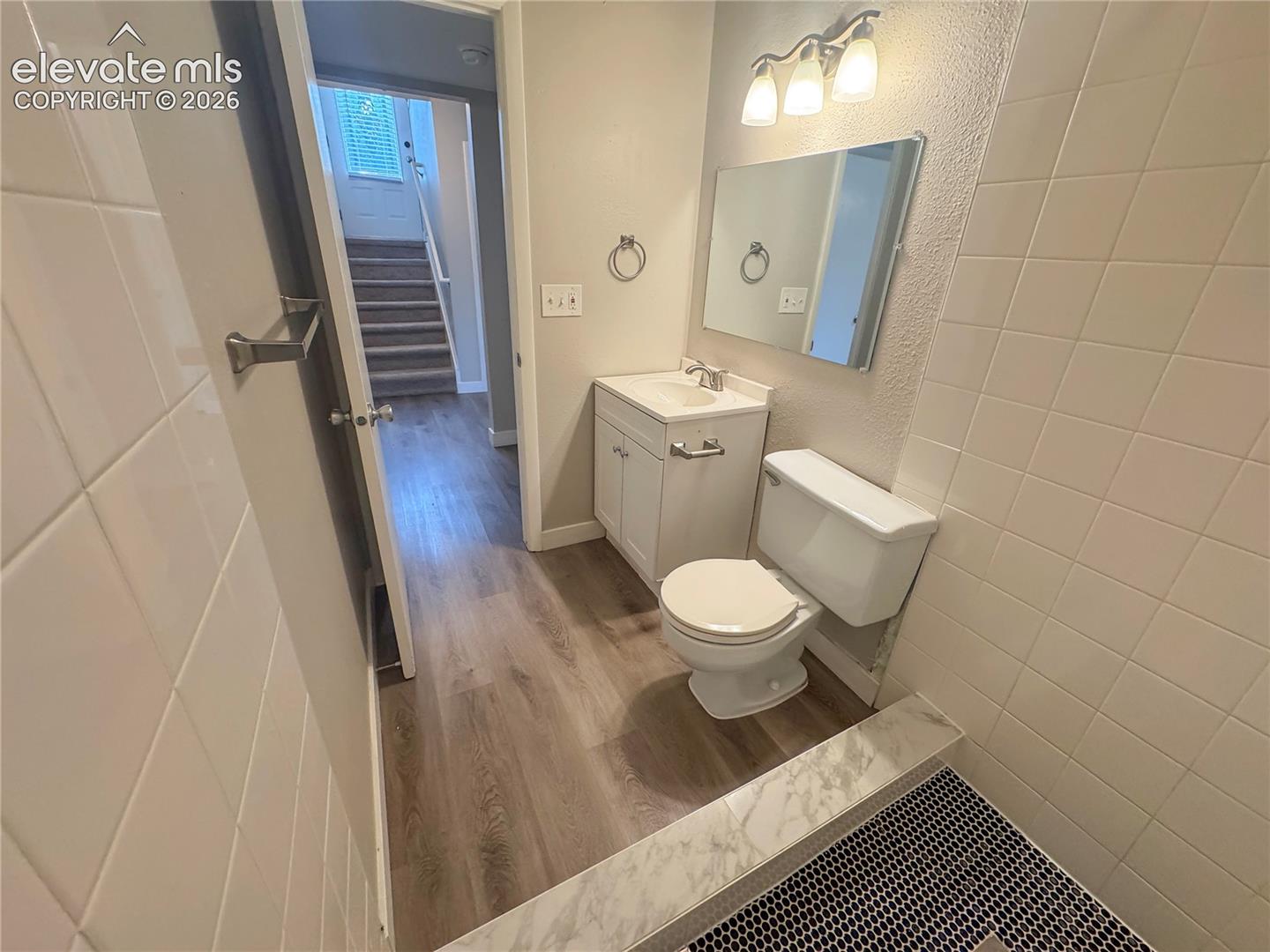 Bathroom featuring wood-finish flooring, white vanity with integrated sink, rectangular wall-mounted mirror, three-light fixture, and tiled shower surround