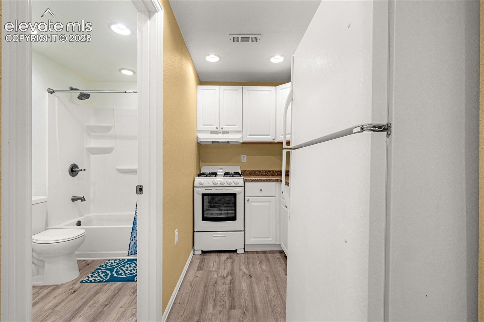 Kitchen featuring white cabinetry, solid-surface countertops, and a white gas range, paired with a bathroom containing a white vanity, a composite tub surround with integrated shelving, and wood-finish flooring throughout