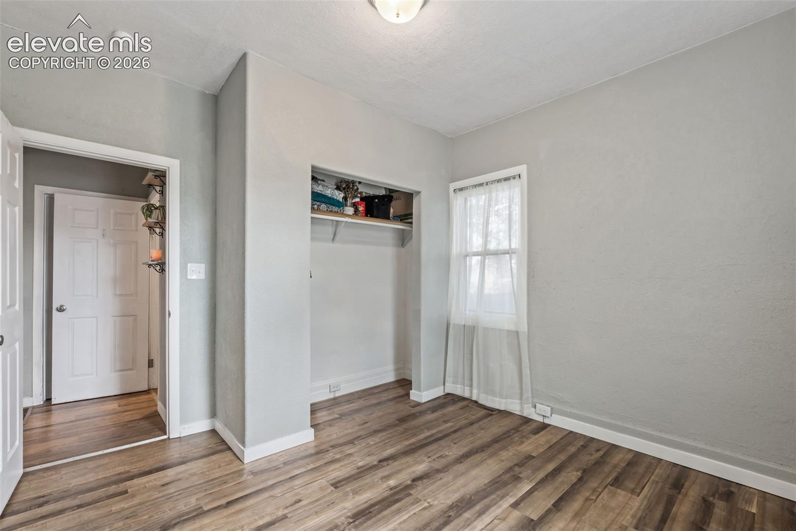 Room featuring wood-finish flooring, light grey painted walls, white trim, and a window with sheer white curtains