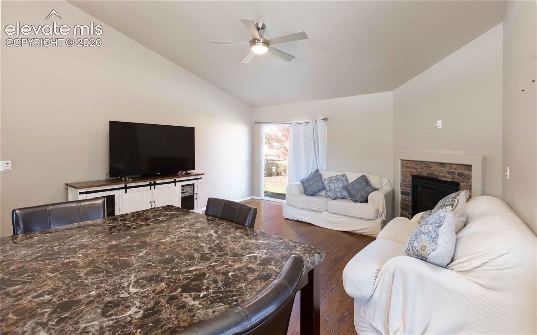 Vaulted ceilings featuring a central overhead fan, stone-surround fireplace, wood-finish flooring, and sliding glass door