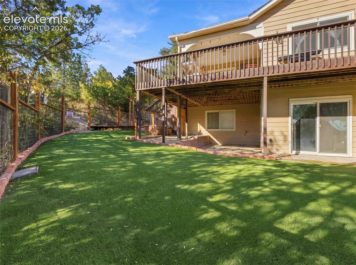 Backyard featuring synthetic turf, elevated wood deck with railing, sliding glass doors, and perimeter wire fencing with wood posts