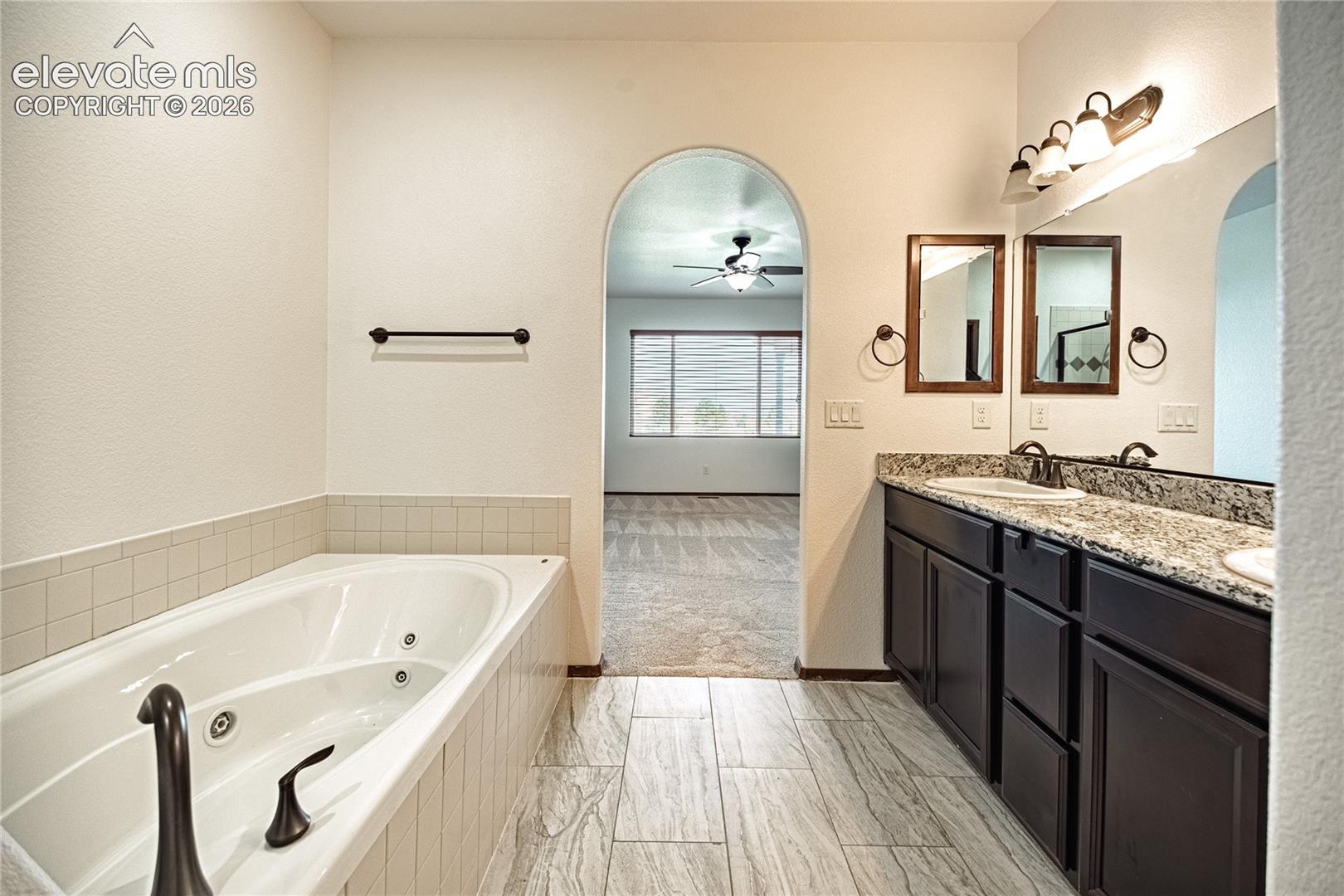 Bathroom featuring a jetted soaking tub, dual vanity with undermount sinks, granite-style countertop, and wood-finish tile flooring