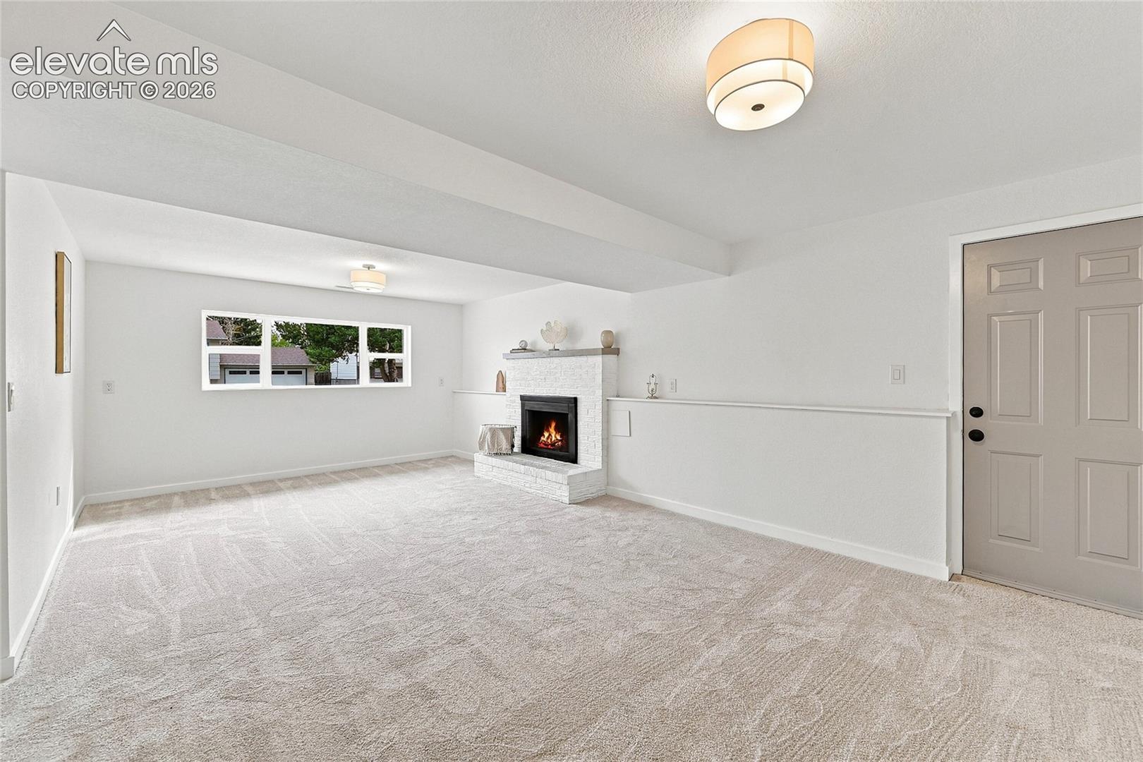 Spacious room featuring light-toned carpeting, a white brick-surround fireplace, and a window with exterior visibility