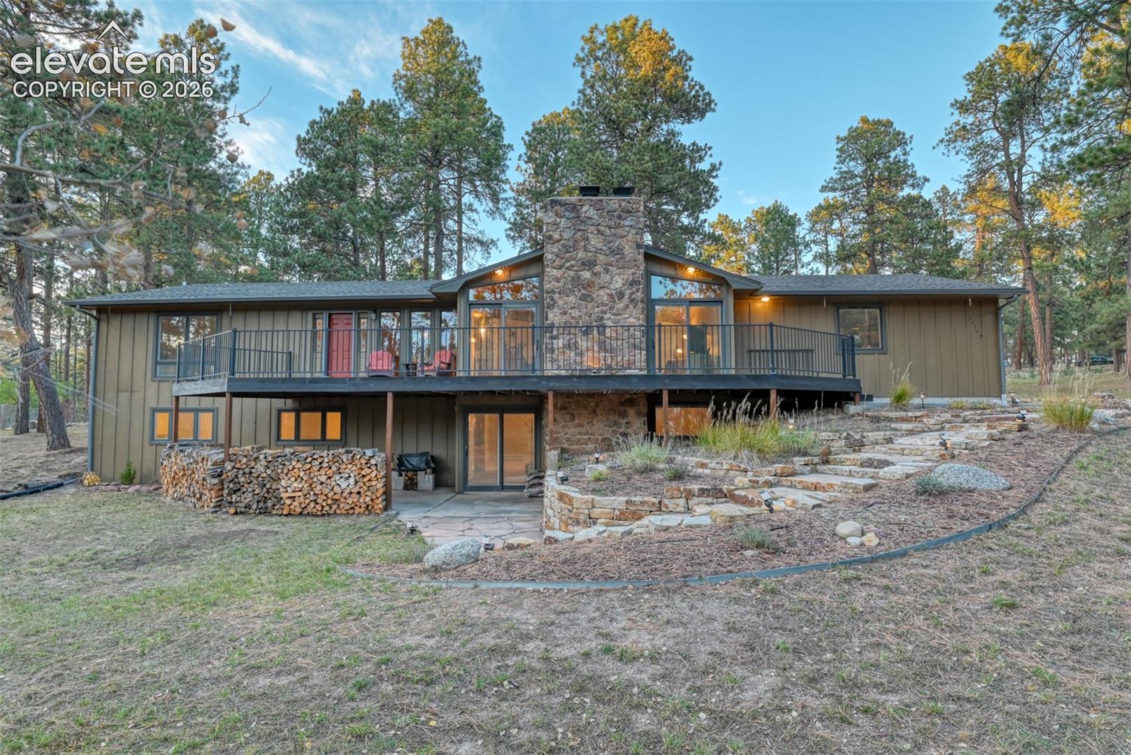 Walk-out ranch residence featuring a prominent stone chimney, expansive raised deck with metal railings, and multi-tiered stone landscaping