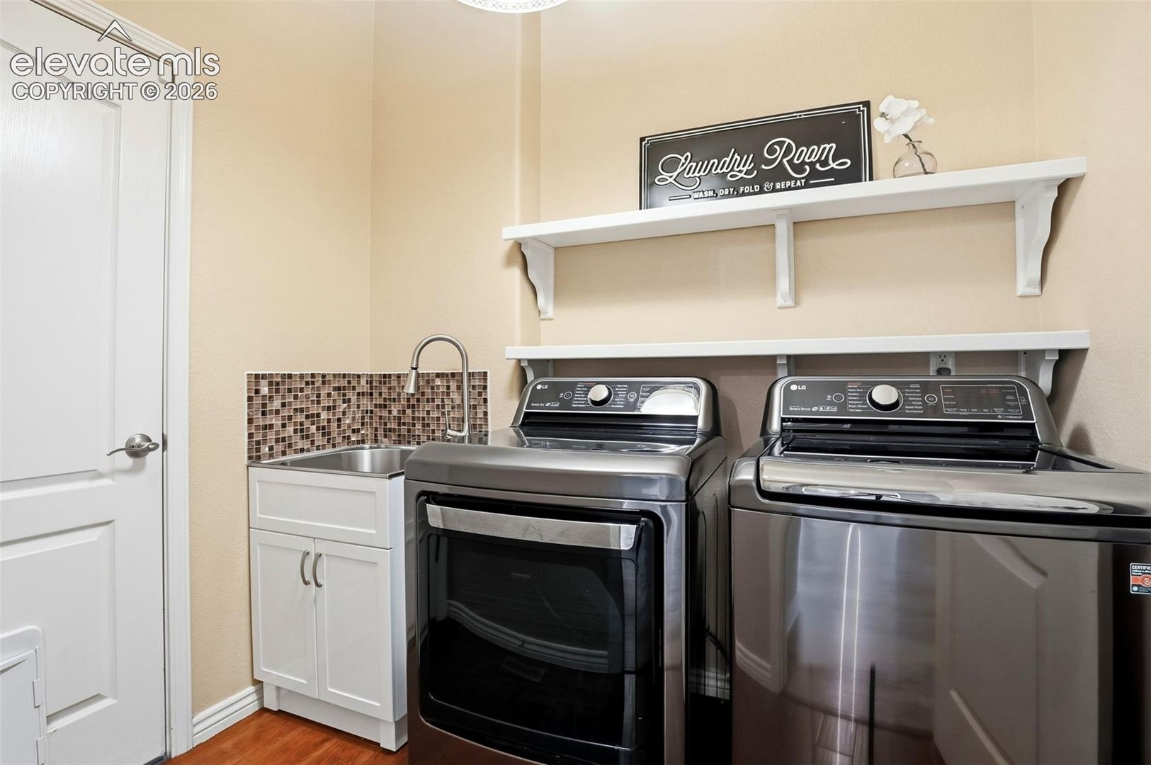Laundry room featuring front-load washer and dryer which are included, mosaic tile backsplash, stainless steel utility sink, and floating shelving for storage and display.