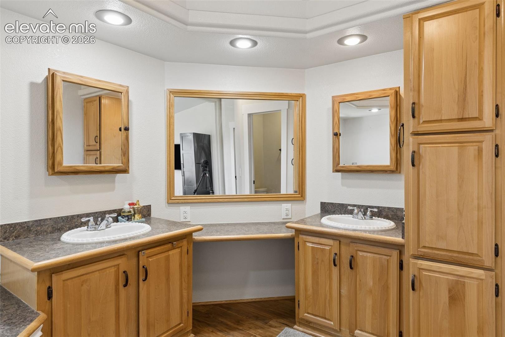 Dual vanity featuring natural wood cabinetry, integrated storage tower, stone-finish countertops, oval drop-in sinks, and framed mirrors