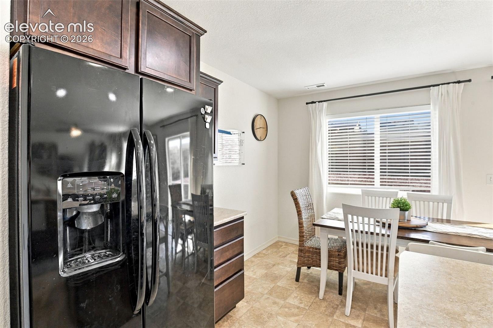 Built-in refrigerator with ice and water dispenser, recessed panel upper cabinetry, light-toned countertop, window with blinds, and patterned tile flooring