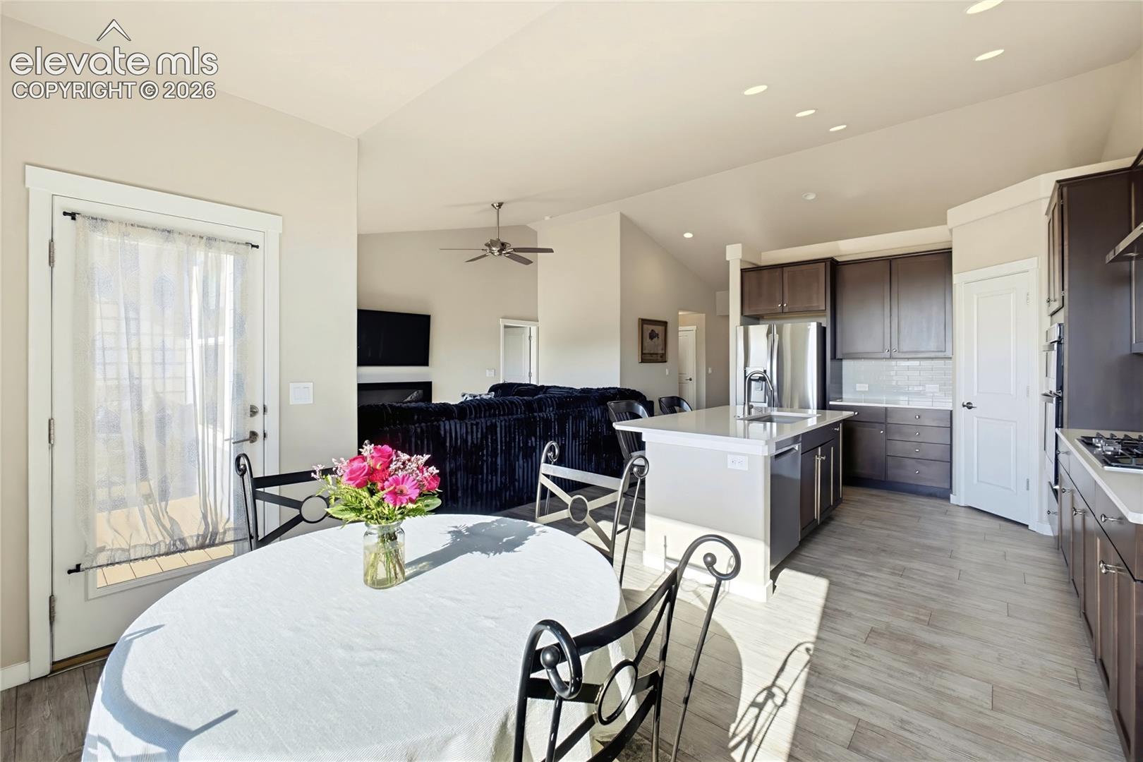 Open-concept kitchen featuring dark-tone cabinetry, stone-surfaced countertops, a center island with integrated sink, and wood-finish flooring under vaulted ceilings with recessed lighting