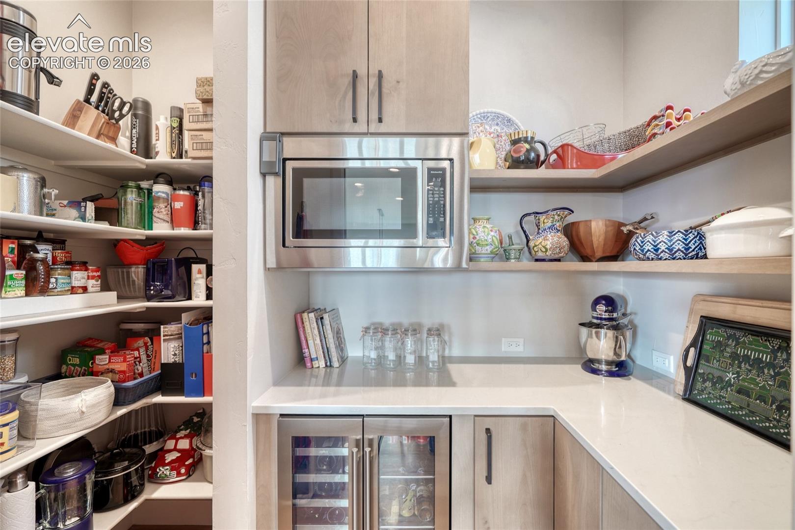 Walk-in pantry featuring light wood-finish cabinetry, stone-style countertops, open shelving, integrated microwave, and built-in under-counter beverage refrigerator