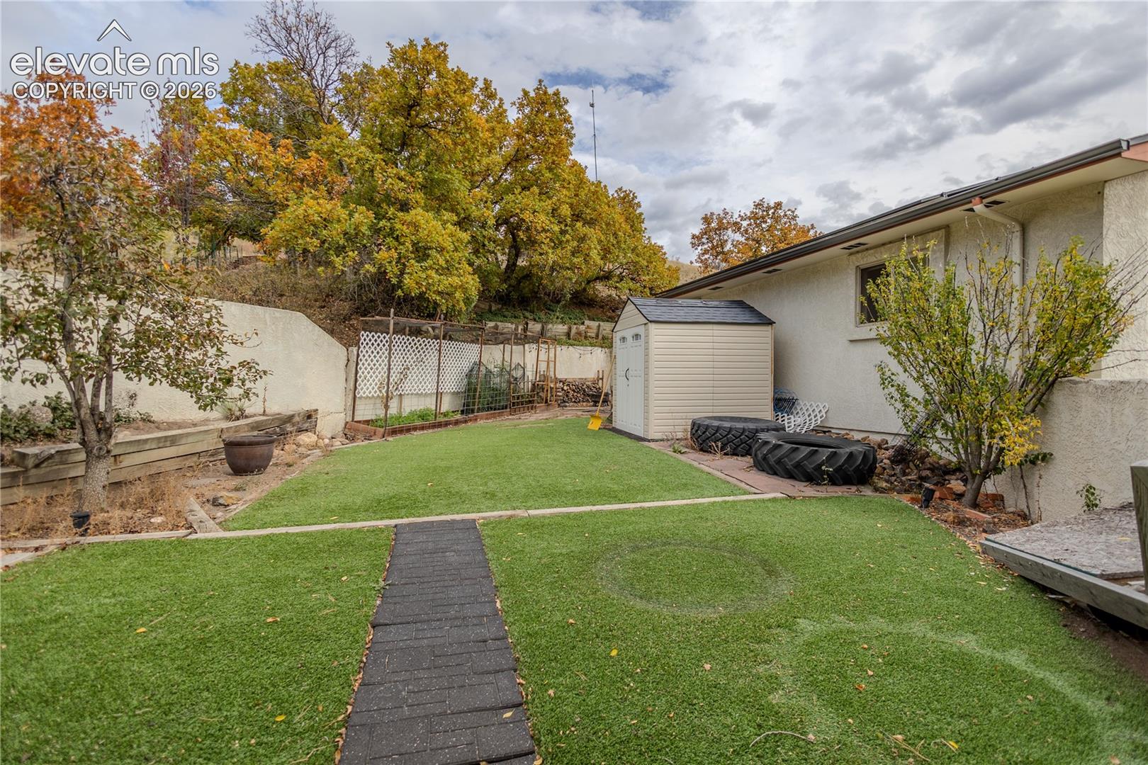 Backyard featuring synthetic turf, retaining wall, built-in trellis, and mature trees
