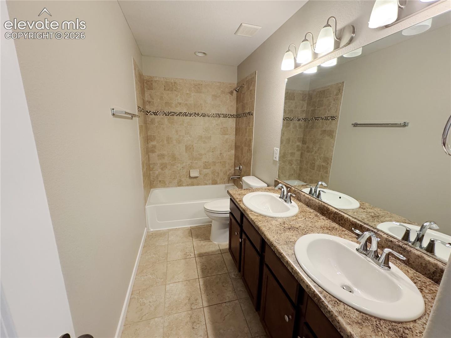 Dual-sink vanity with stone-finish countertop, tile-surround bathtub, ceramic tile flooring, and multi-bulb vanity lighting