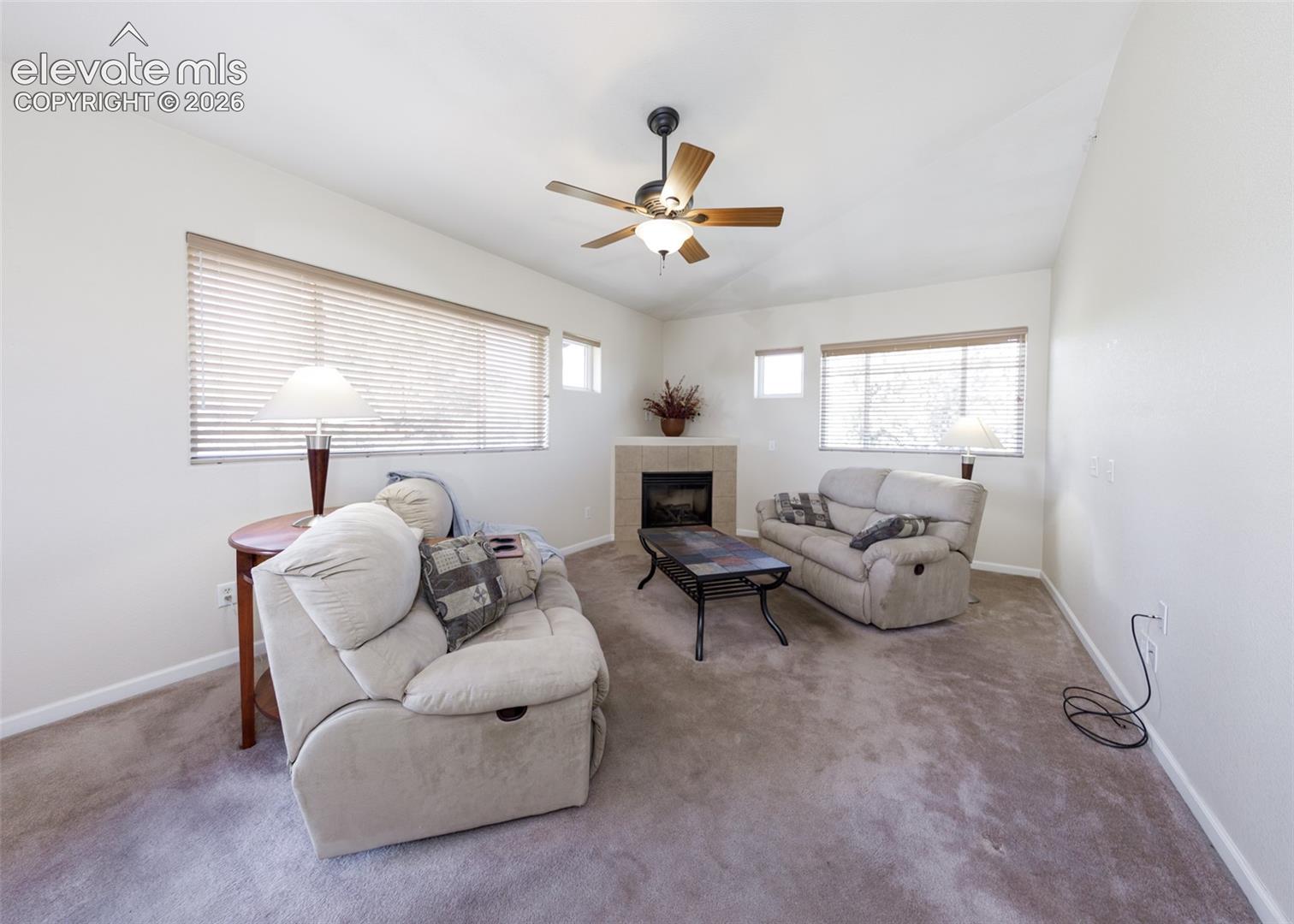 Corner fireplace with tile surround, neutral carpeting, ceiling fan with integrated lighting, and multiple windows with horizontal blinds