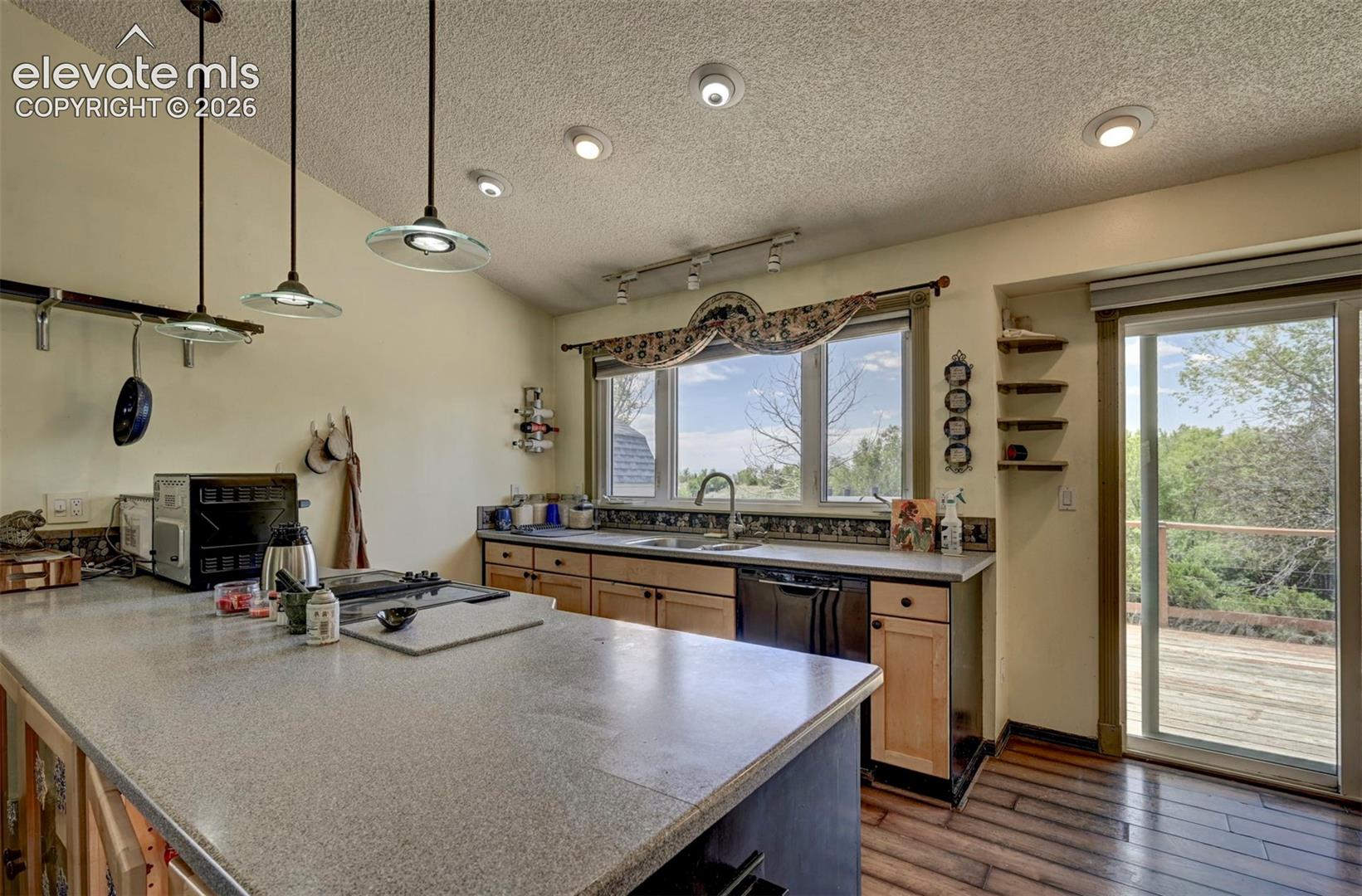 Kitchen featuring a large central island with a speckled countertop, natural wood cabinetry, a sliding glass door providing outdoor access, a black dishwasher, and pendant lighting