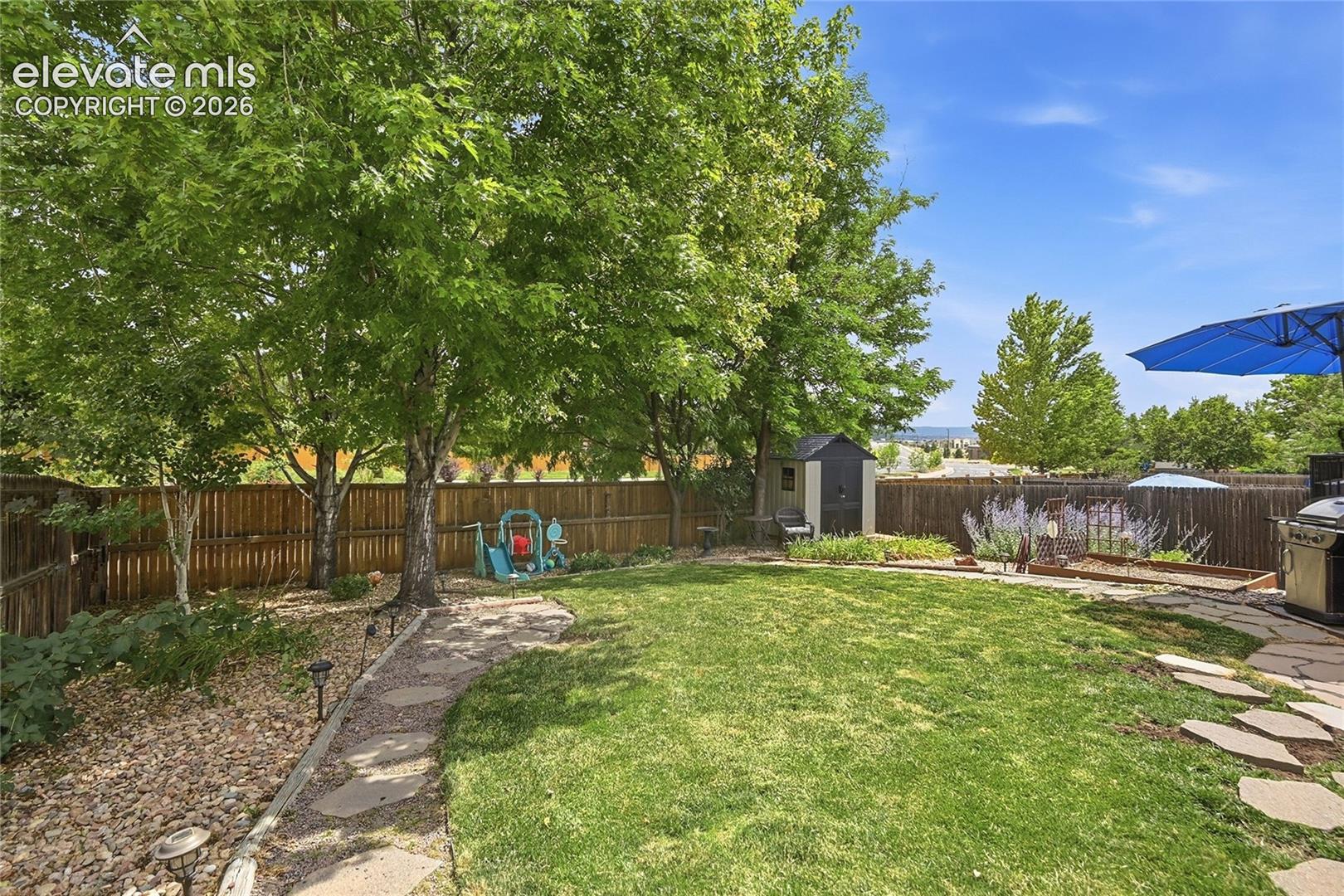 Fenced backyard featuring a manicured lawn, stone paver pathway, mature trees, and a detached storage shed