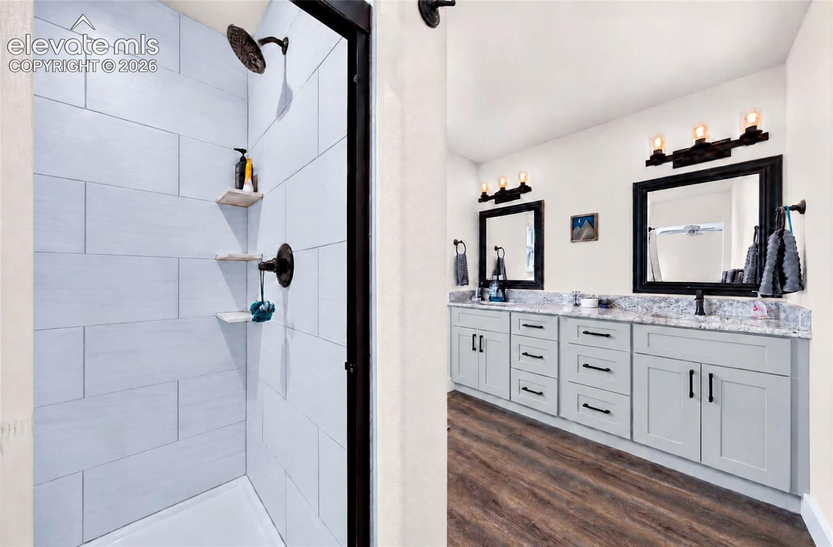 Bathroom featuring a dual-sink vanity with light-toned cabinetry, patterned stone-finish countertops, wood-finish flooring, and a walk-in shower with large-format wall tile