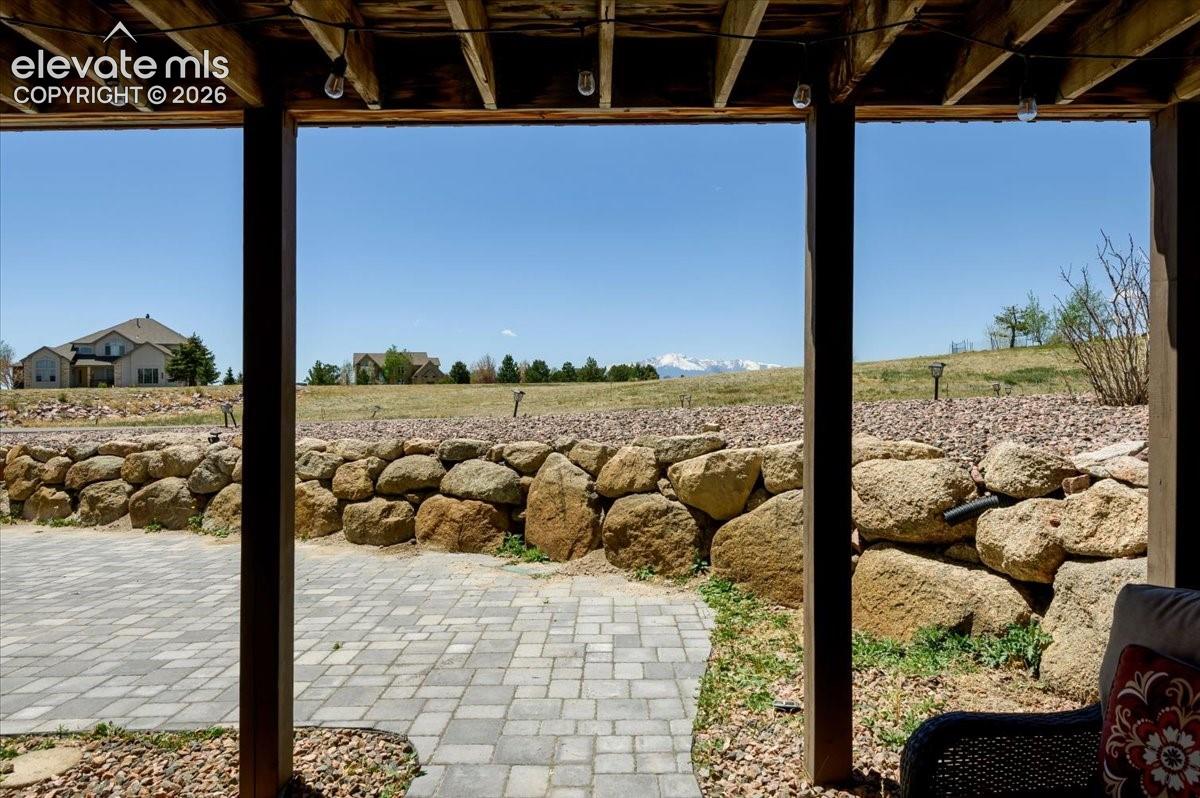 Covered patio space featuring an exposed wood joist ceiling with string lighting, a paver patio, and a natural stone retaining wall