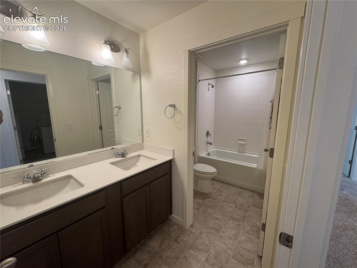 Dual-sink vanity with light-toned solid surface countertop, dark wood-finish cabinetry, full-width mirror, and tiled shower-tub combination