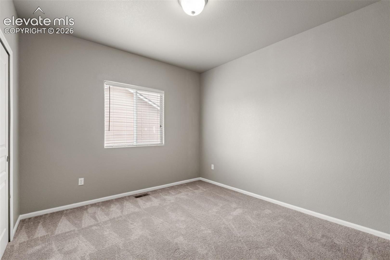 Neutral-toned interior space featuring textured wall-to-wall carpeting, a single window with horizontal blinds, and a dome-style flush mount ceiling light