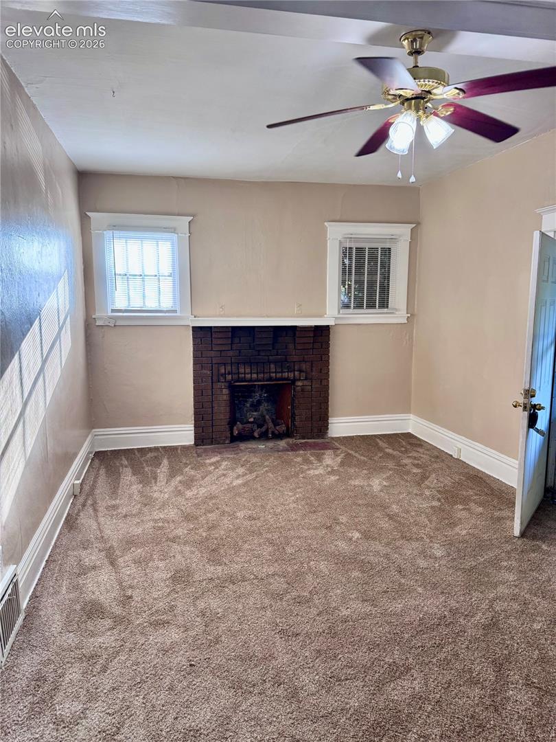 Brick fireplace feature, two windows with grilles, ceiling fan with integrated lighting, and wall-to-wall carpeting