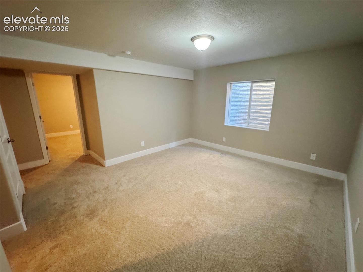 Neutral-toned interior space featuring wall-to-wall carpeting, an inset window with horizontal blinds, and a central ceiling-mounted light fixture