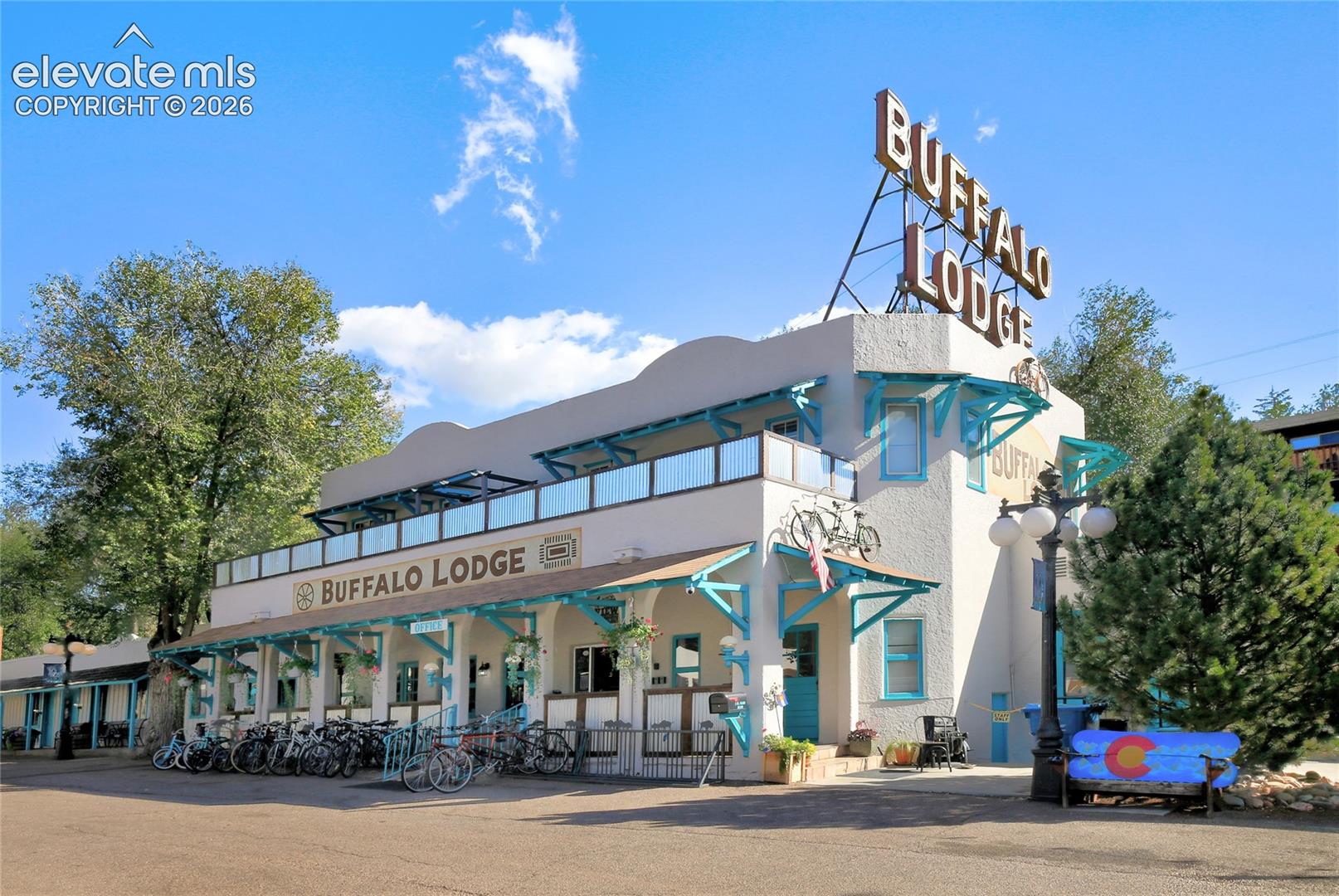 Two-story stucco exterior featuring a covered front porch, teal structural brackets, balcony with glass railing, and decorative rooftop signage