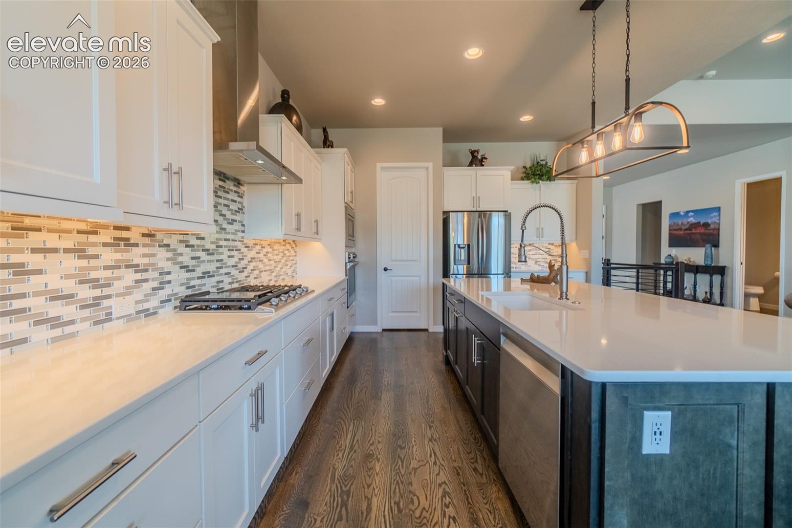 Spacious kitchen featuring an expansive island with an undermount sink, stainless steel appliances, white cabinetry, a glass tile backsplash, and wood-finish flooring