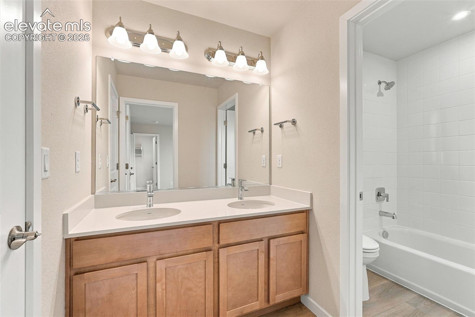 Dual-sink vanity with a solid-surface countertop and wood-finish cabinetry, complemented by wall-mounted lighting and a combination tub and shower enclosure with white tiled surround