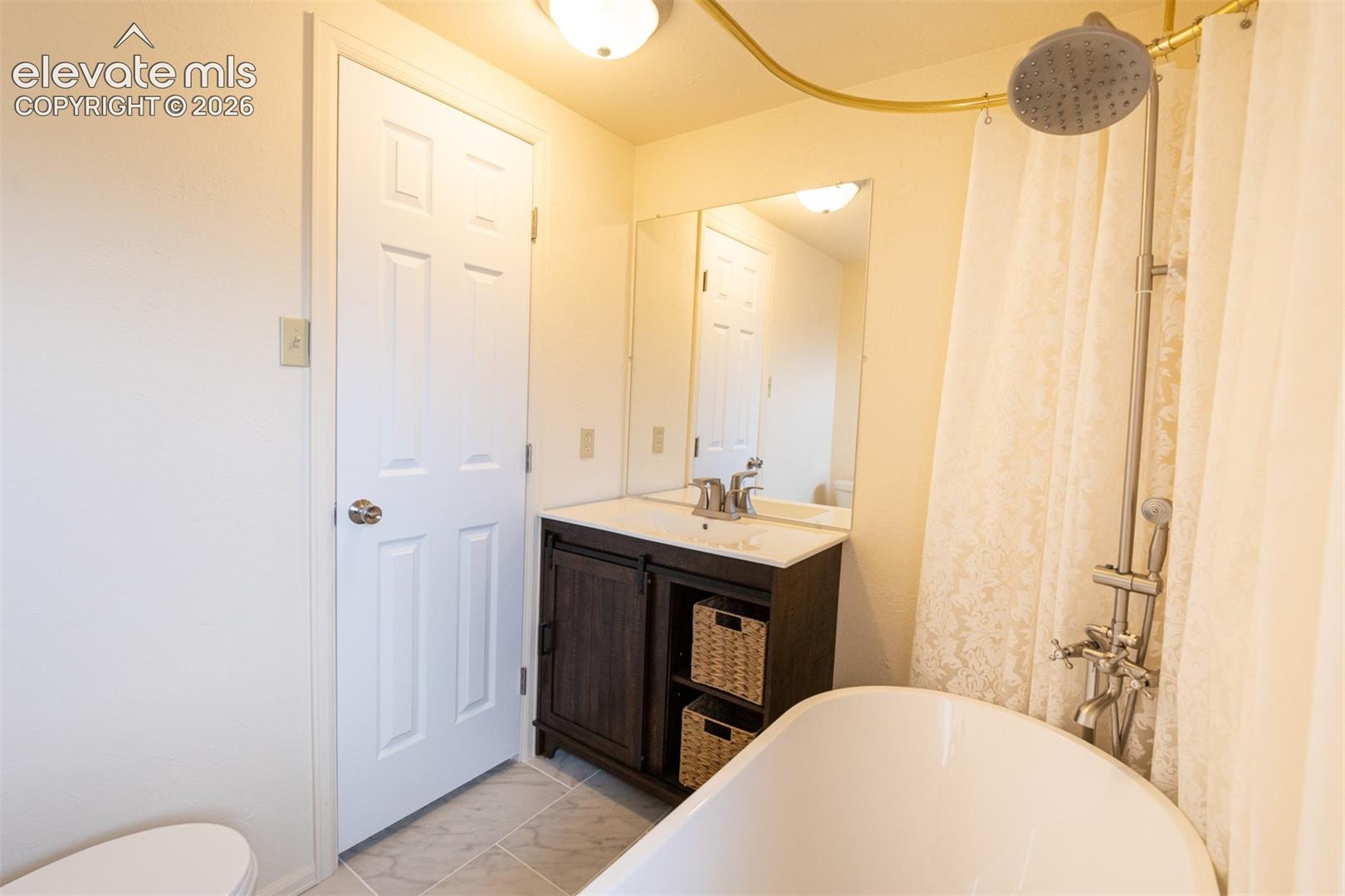Bathroom featuring a freestanding tub with a metallic shower assembly, wood-finish vanity with open shelving, neutral-tone tiled flooring, and a wall-mounted mirror