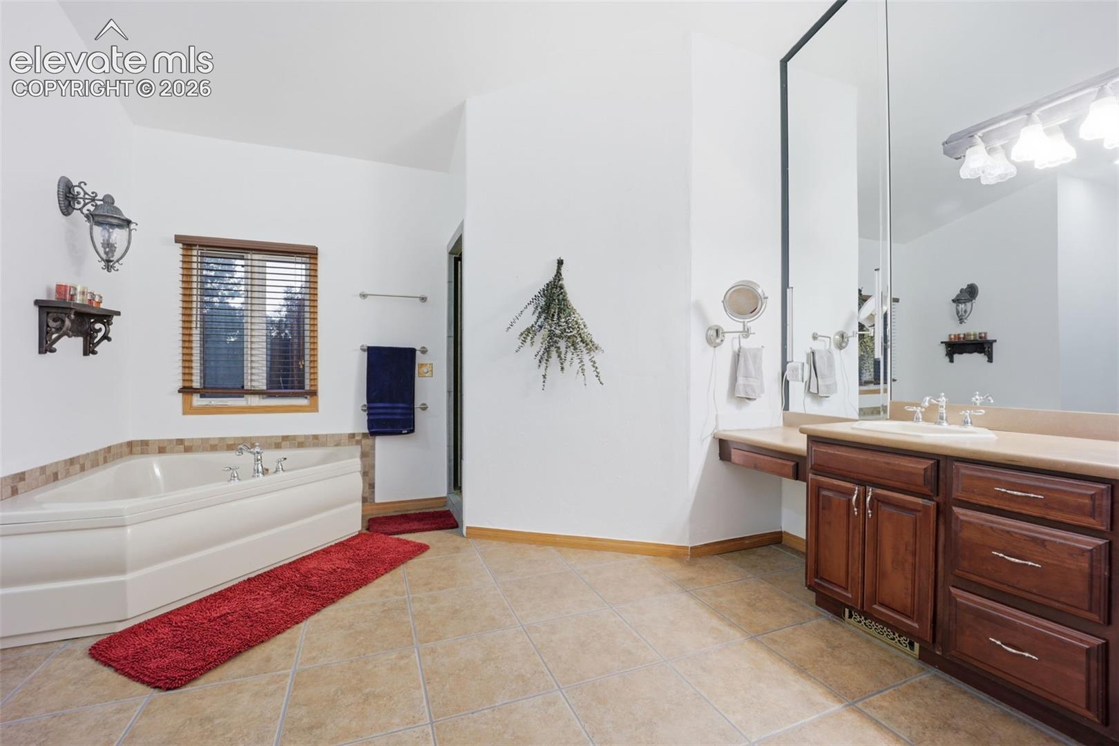 Tile flooring, built-in soaking tub with tile surround, wood-finish vanity with integrated sink, spacious mirror, and mounted vanity lighting