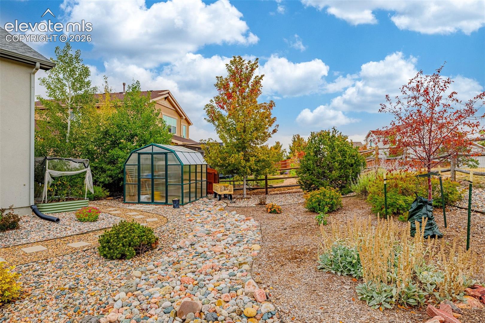 Xeriscape backyard featuring a greenhouse, stone pathways, varied plantings, and timber fencing