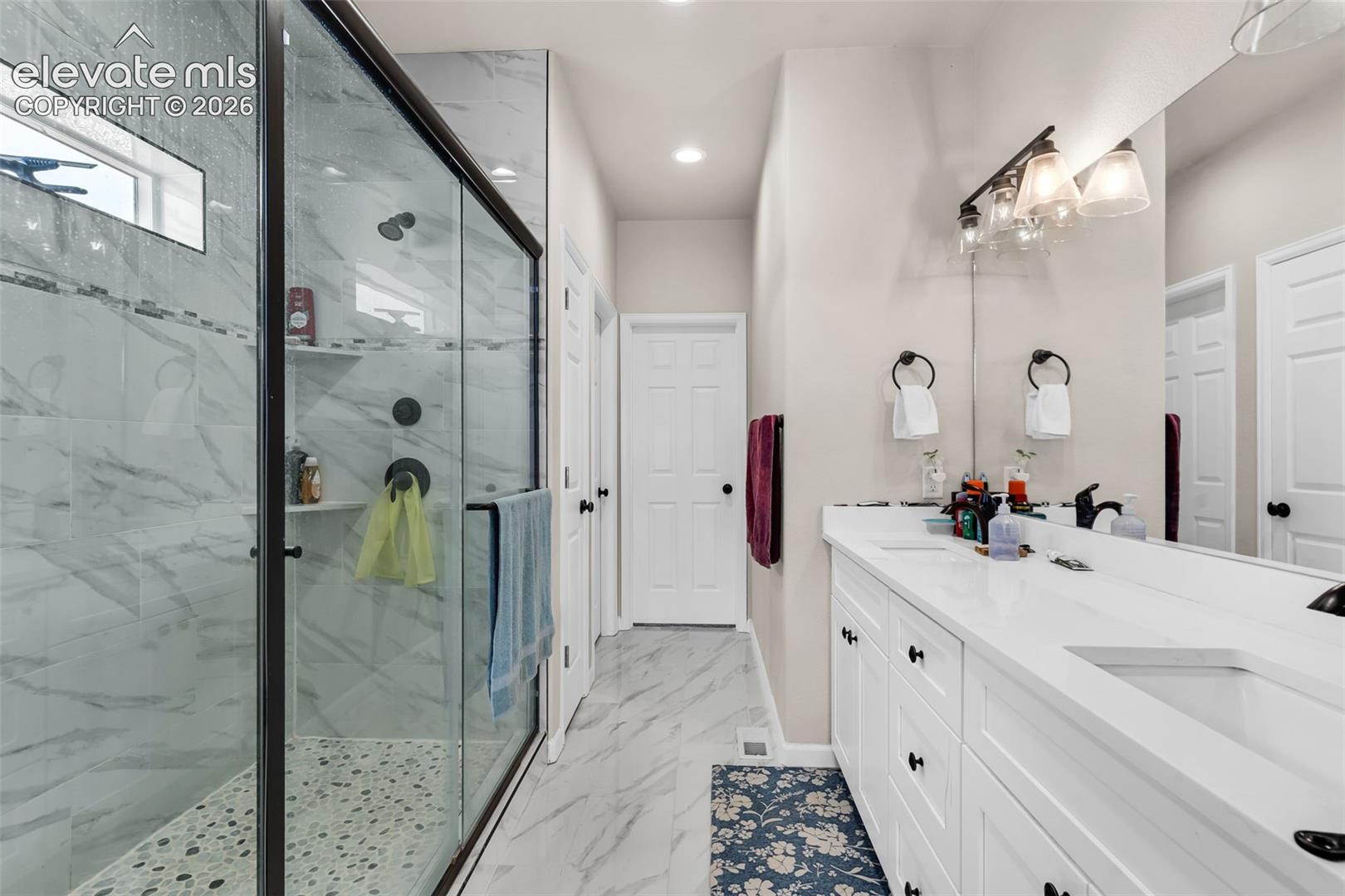 Spacious bathroom featuring a frameless glass shower enclosure with a pebble-stone floor and built-in shelving, dual vanity with undermount sinks, stone countertops, and a full-width mirror