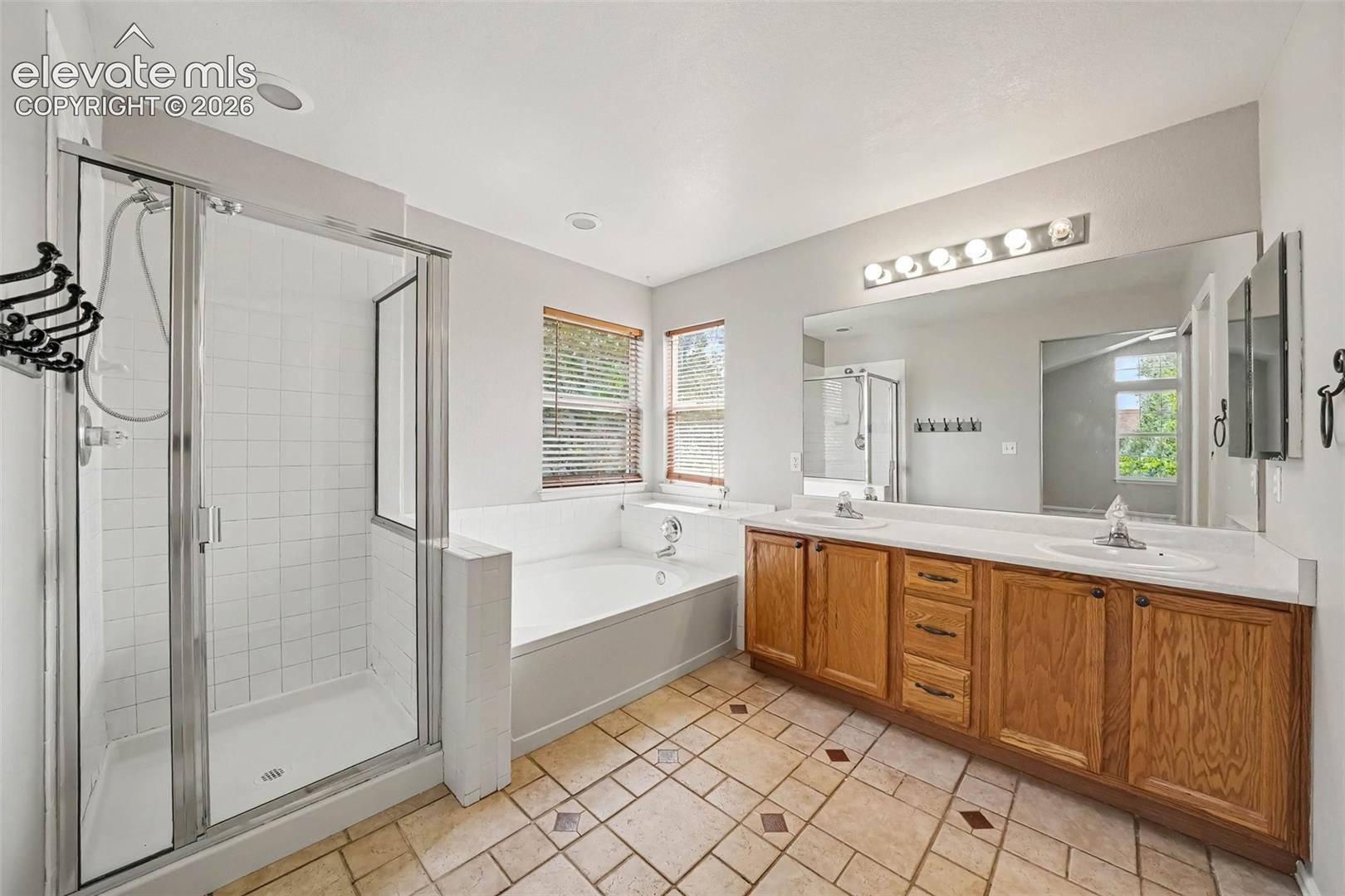 Dual vanity with integrated sinks, full-width mirror, white tile flooring, enclosed shower with a glass door, and a built-in bathtub