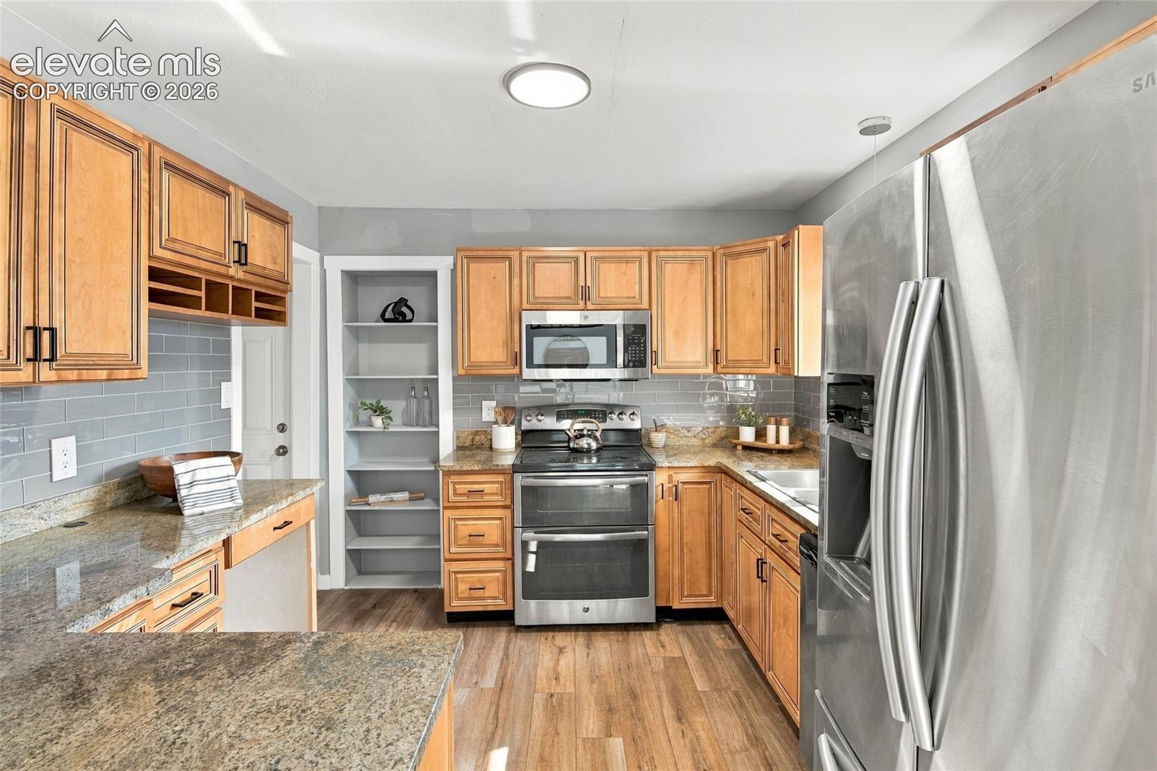 Kitchen featuring shaker-style wood cabinetry, stone countertops, gray subway tile backsplash, stainless steel appliances, and built-in shelving