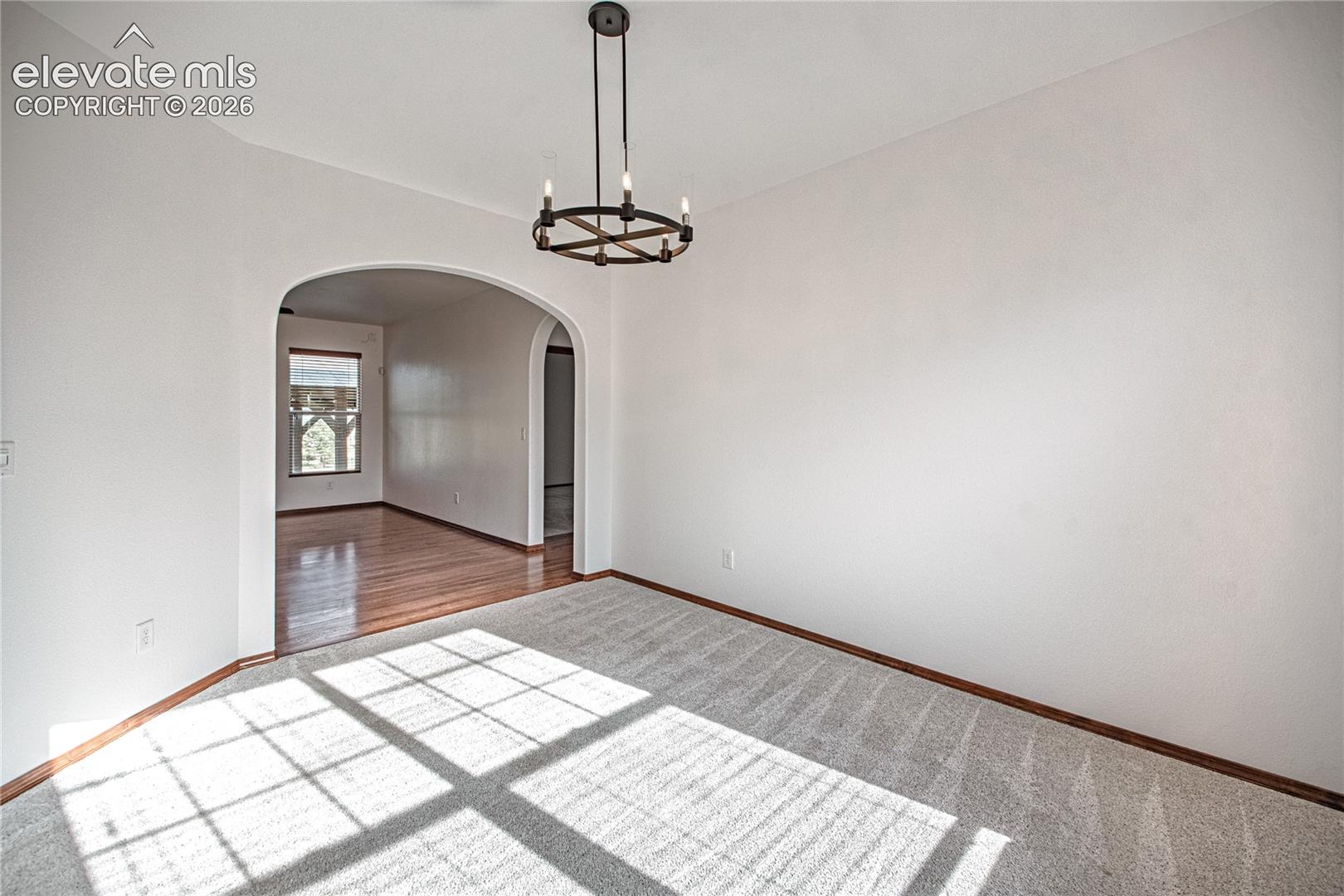 Room featuring light-colored carpeting, a dark bronze-finish chandelier, and an arched doorway opening to a room with wood flooring