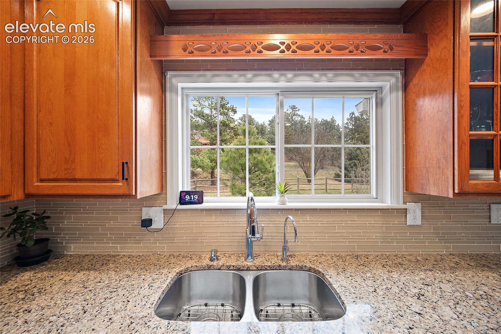 Kitchen featuring wood cabinetry, decorative millwork, stone-finish countertops, linear tile backsplash, and a double-basin stainless steel sink under a multi-pane window
