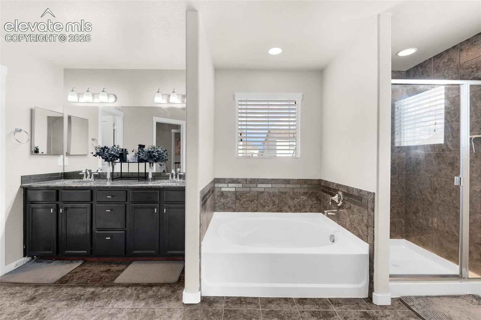 Bathroom featuring a dual-sink dark wood vanity with stone-style countertop, recessed lighting, soaking tub with tile surround, and a glass-enclosed shower