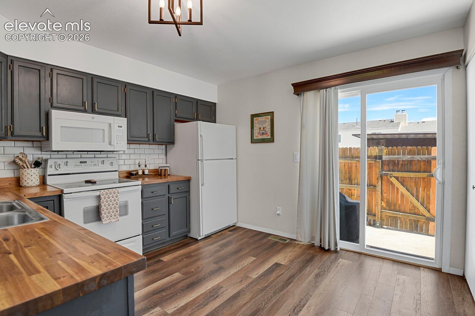 White appliances, wood-finish flooring, dark gray cabinetry with gold hardware, white subway tile backsplash, and a sliding glass door
