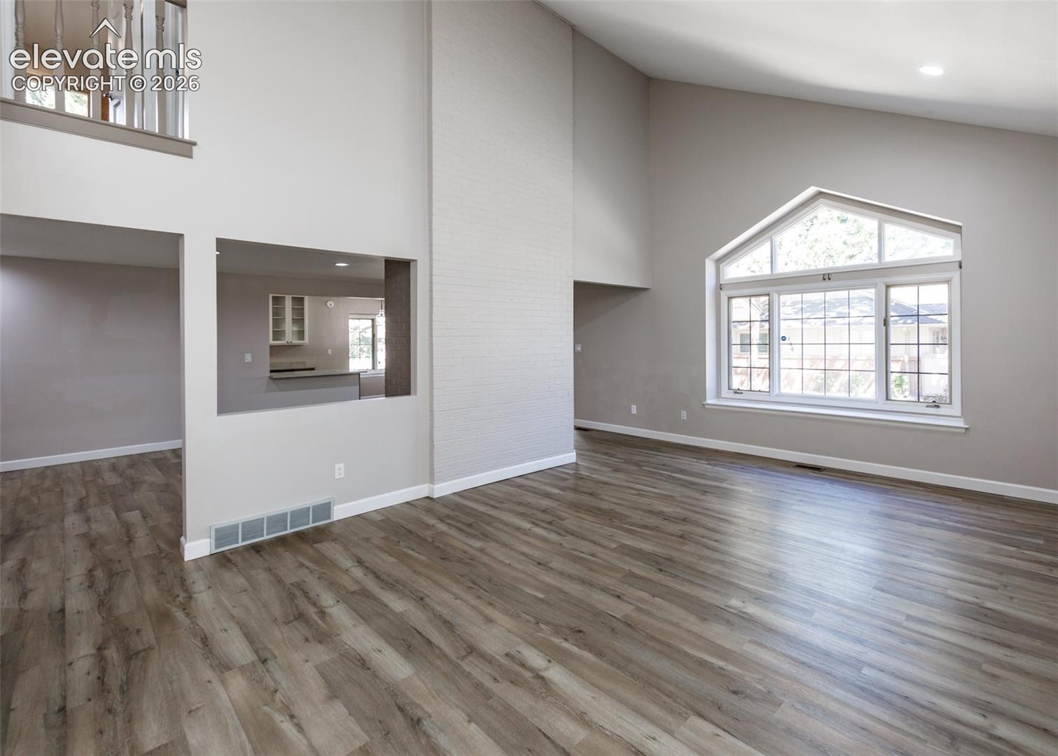 Vaulted ceilings, wood-finish flooring, a large arched window with grid details, a mezzanine balcony, and a pass-through window to the kitchen