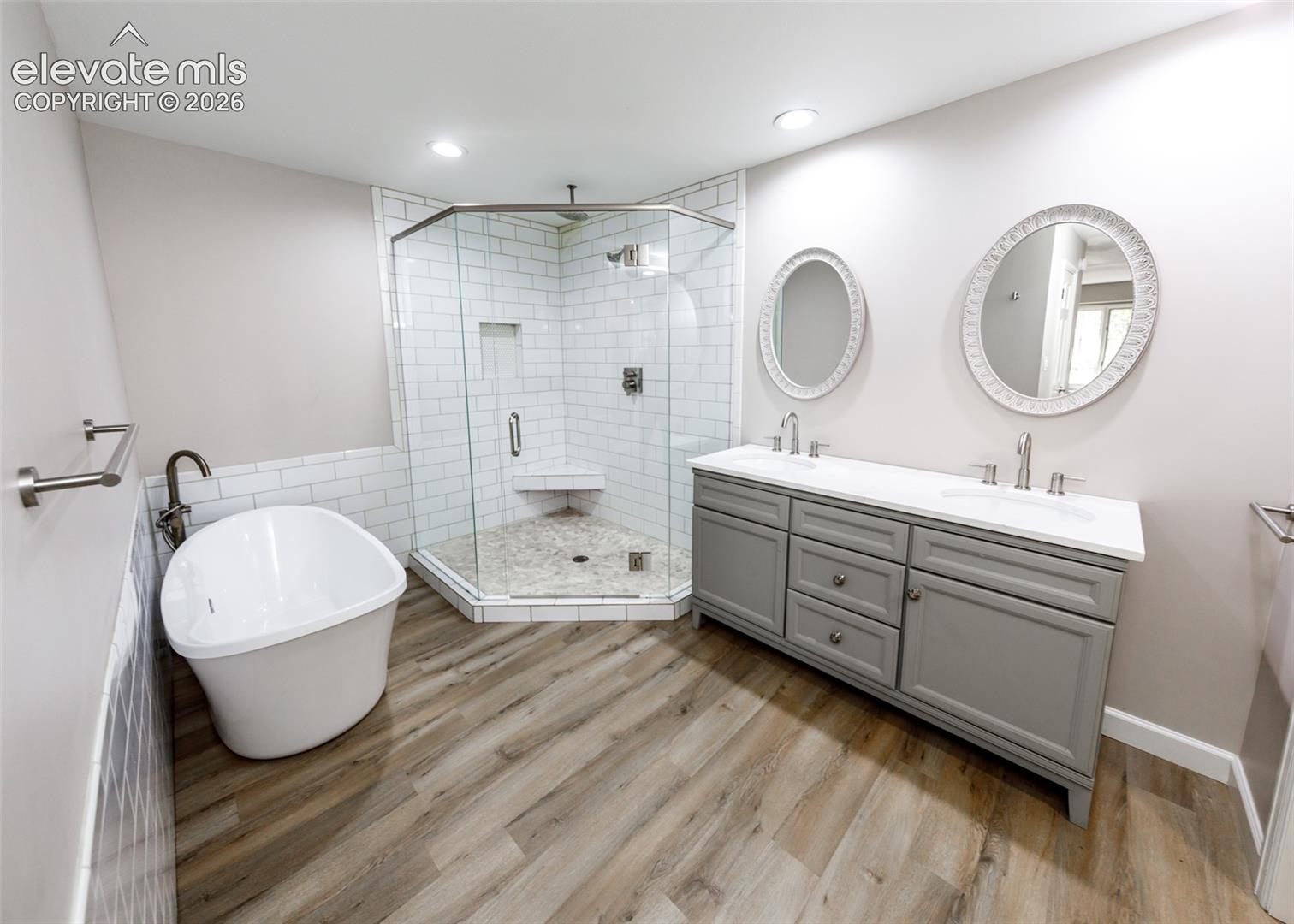 Freestanding soaking tub, wood-finish flooring, glass-enclosed shower with subway tile, dual vanity with undermount sinks, and recessed lighting