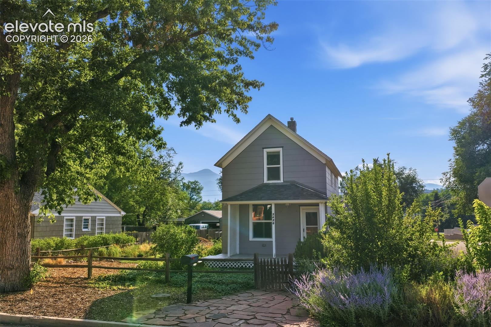 Gabled roofline with a chimney, gray siding exterior, covered front porch, stone pathway, and lush landscaping.