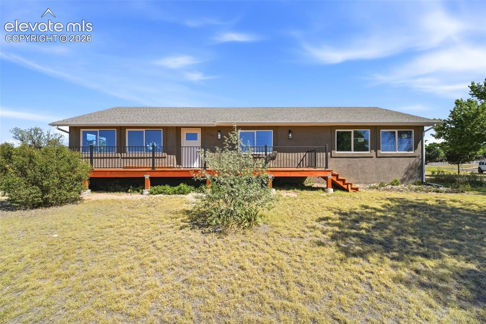 Ranch-style residence featuring a stucco exterior, a neutral-toned roof, a front wood-finish deck with dark railings, and numerous windows