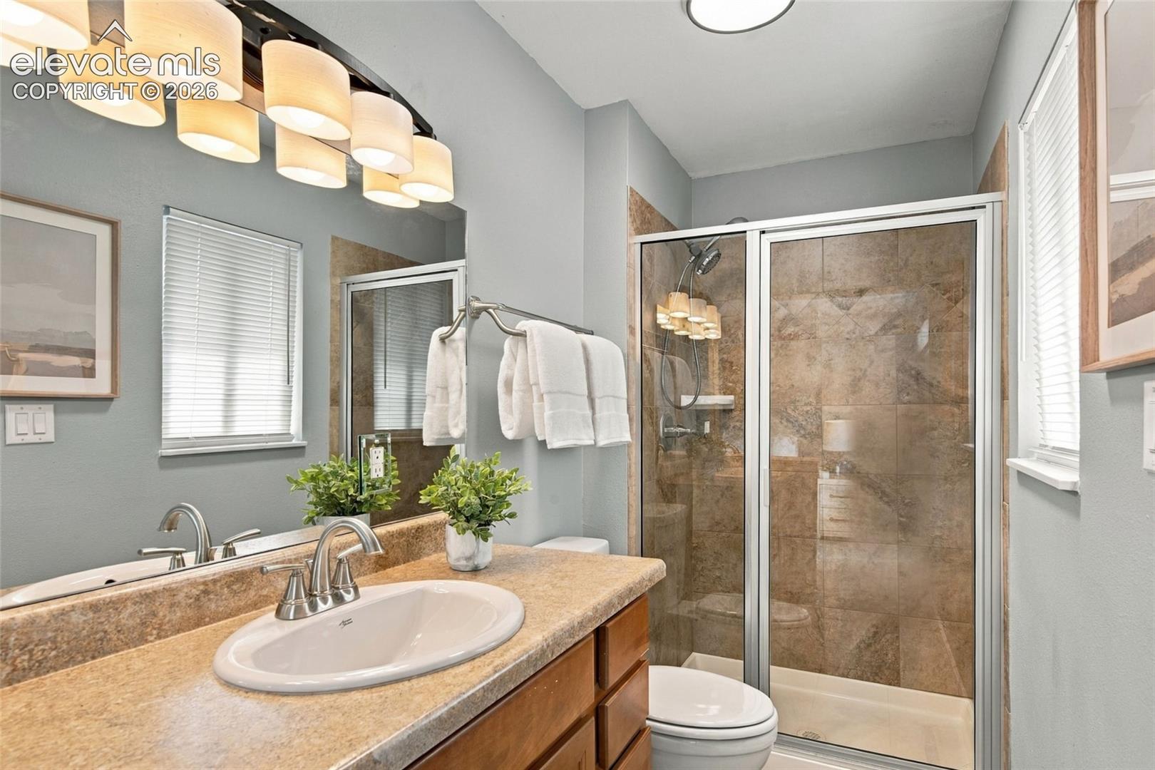 Bathroom featuring an integrated oval sink with a metallic faucet, solid-surface vanity countertop, multi-light vanity fixture, and a glass-enclosed shower with tile surround