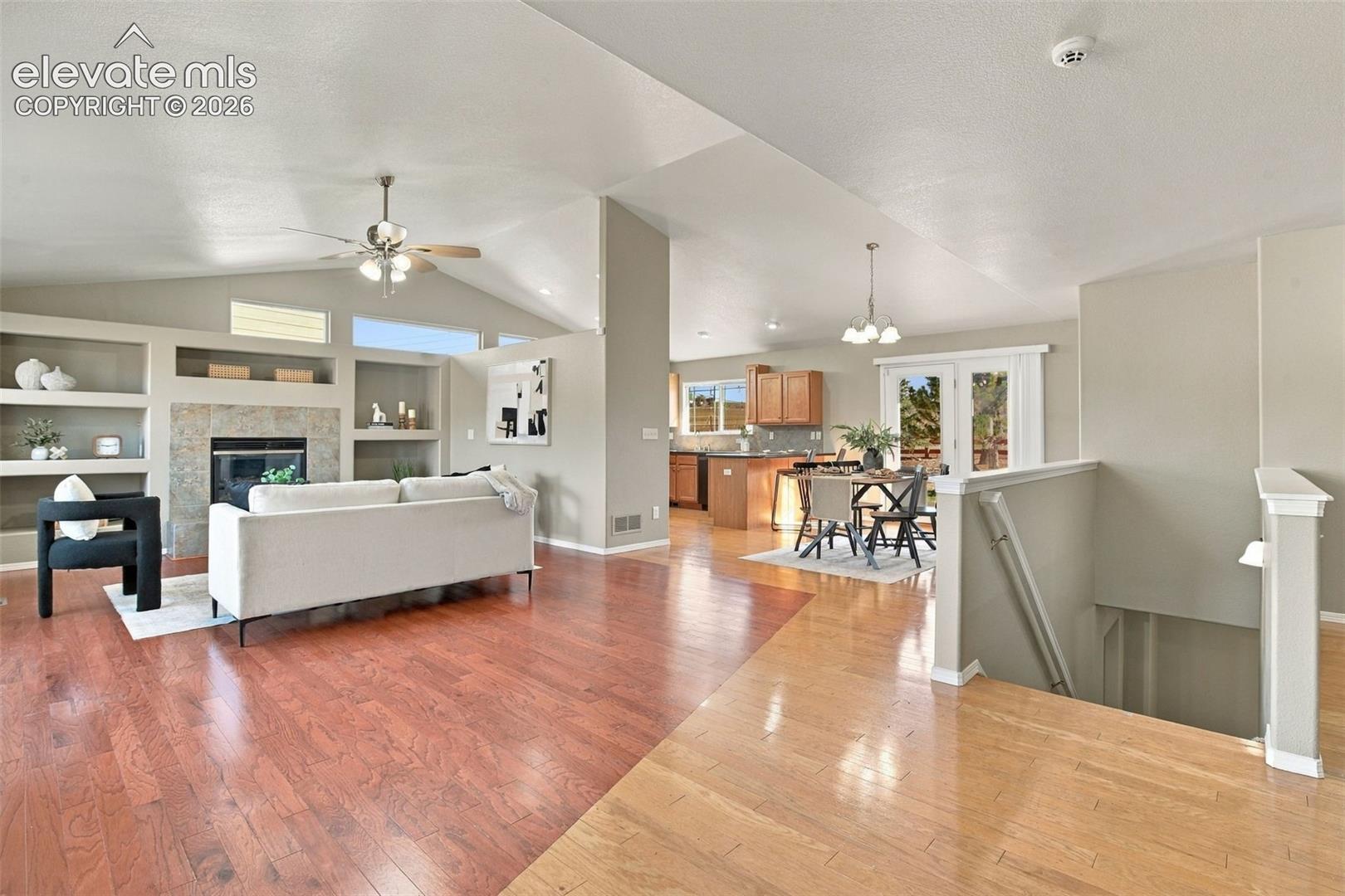 Vaulted ceilings with a ceiling fan, built-in shelving, a fireplace with stone surround, wood-finish flooring, and a kitchen with wood cabinetry