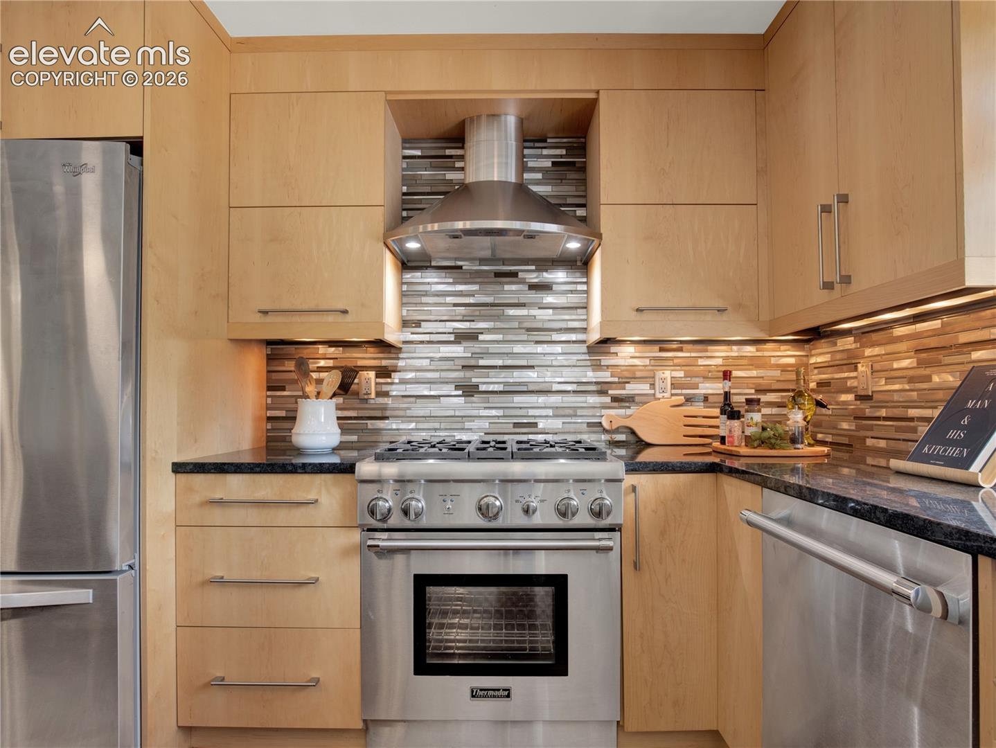 Custom light-toned cabinetry, stainless steel Thermador gas range, integrated range hood, mosaic tile backsplash, and dark stone countertops