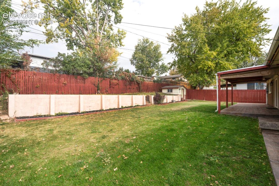 Backyard featuring a covered patio, tiered concrete block retaining wall, mature trees, and perimeter privacy fencing