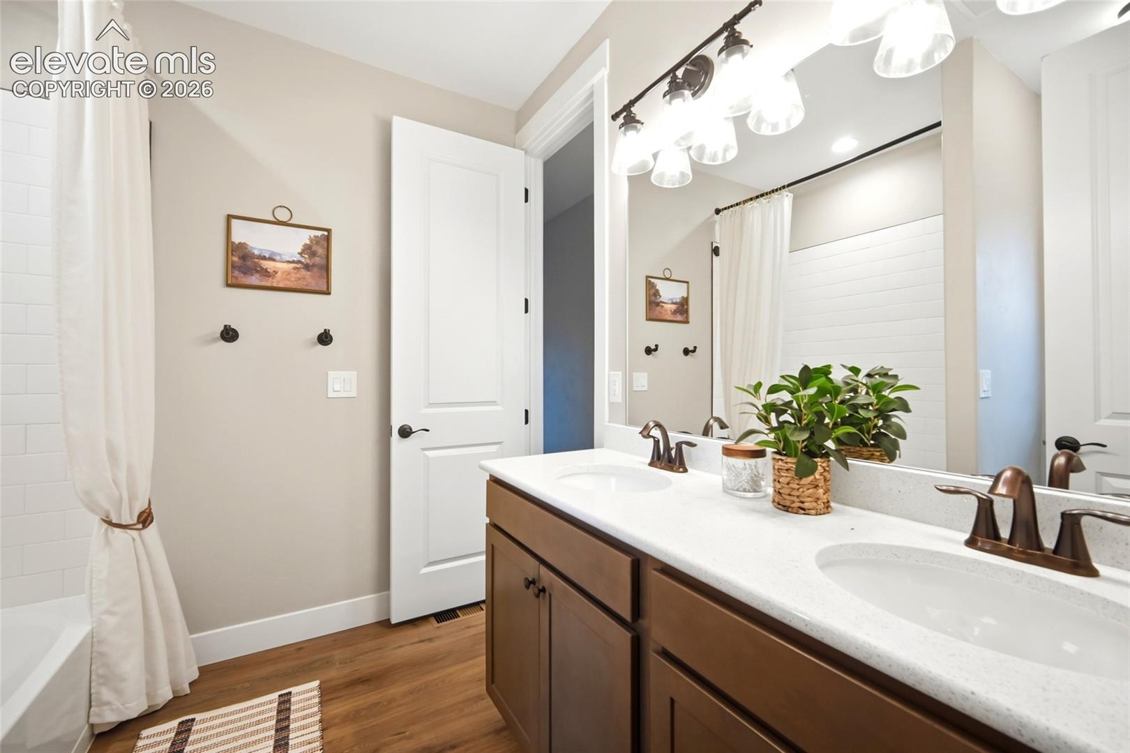 Bathroom featuring a dual-sink vanity with a light-toned solid surface countertop, dark-finish hardware, wood-finish flooring, and a full-width vanity mirror with integrated lighting