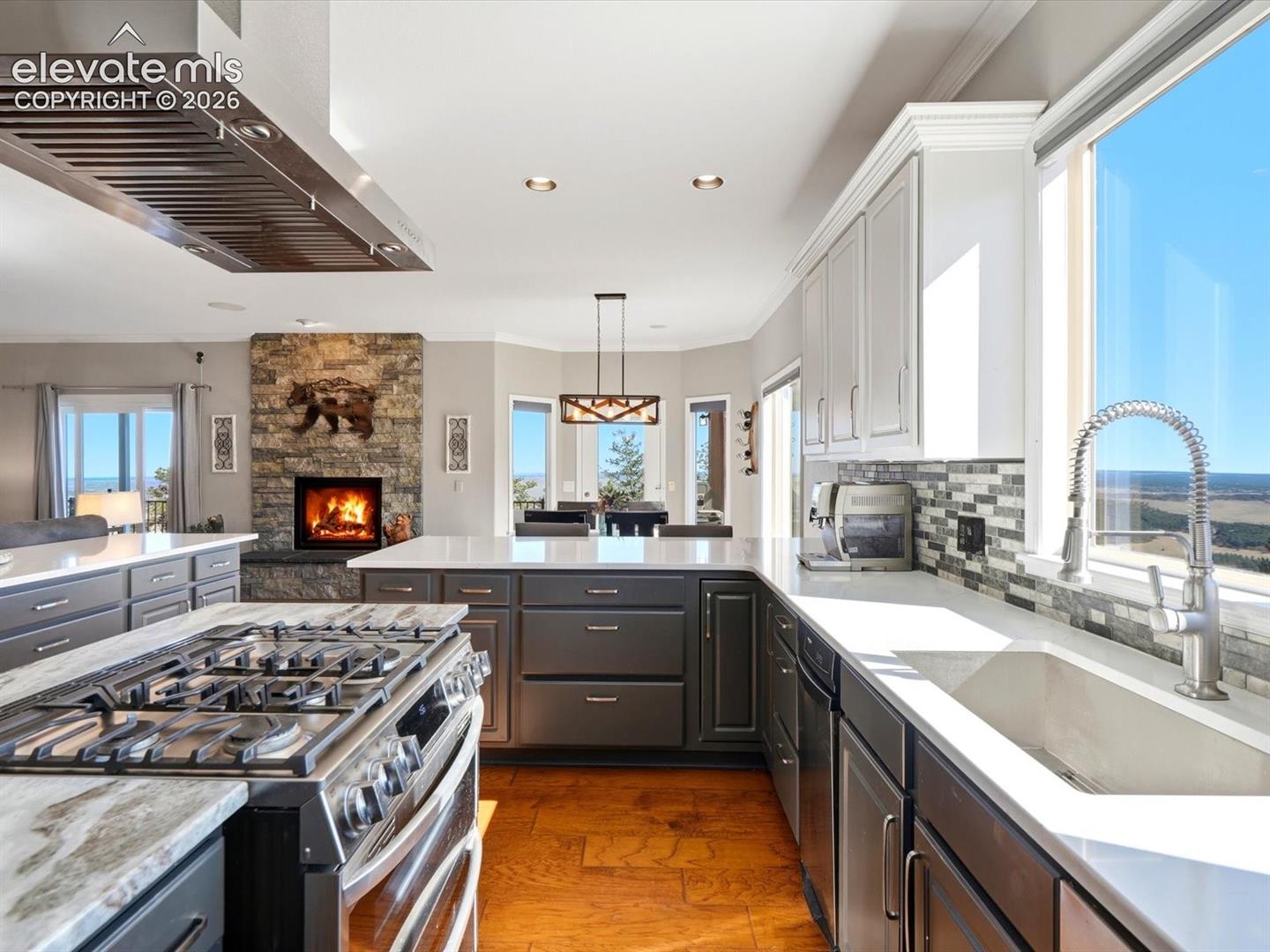 Kitchen featuring quartz countertops, dark cabinetry, a professional-grade gas range with stainless steel hood, mosaic tile backsplash, and stone-surround fireplace