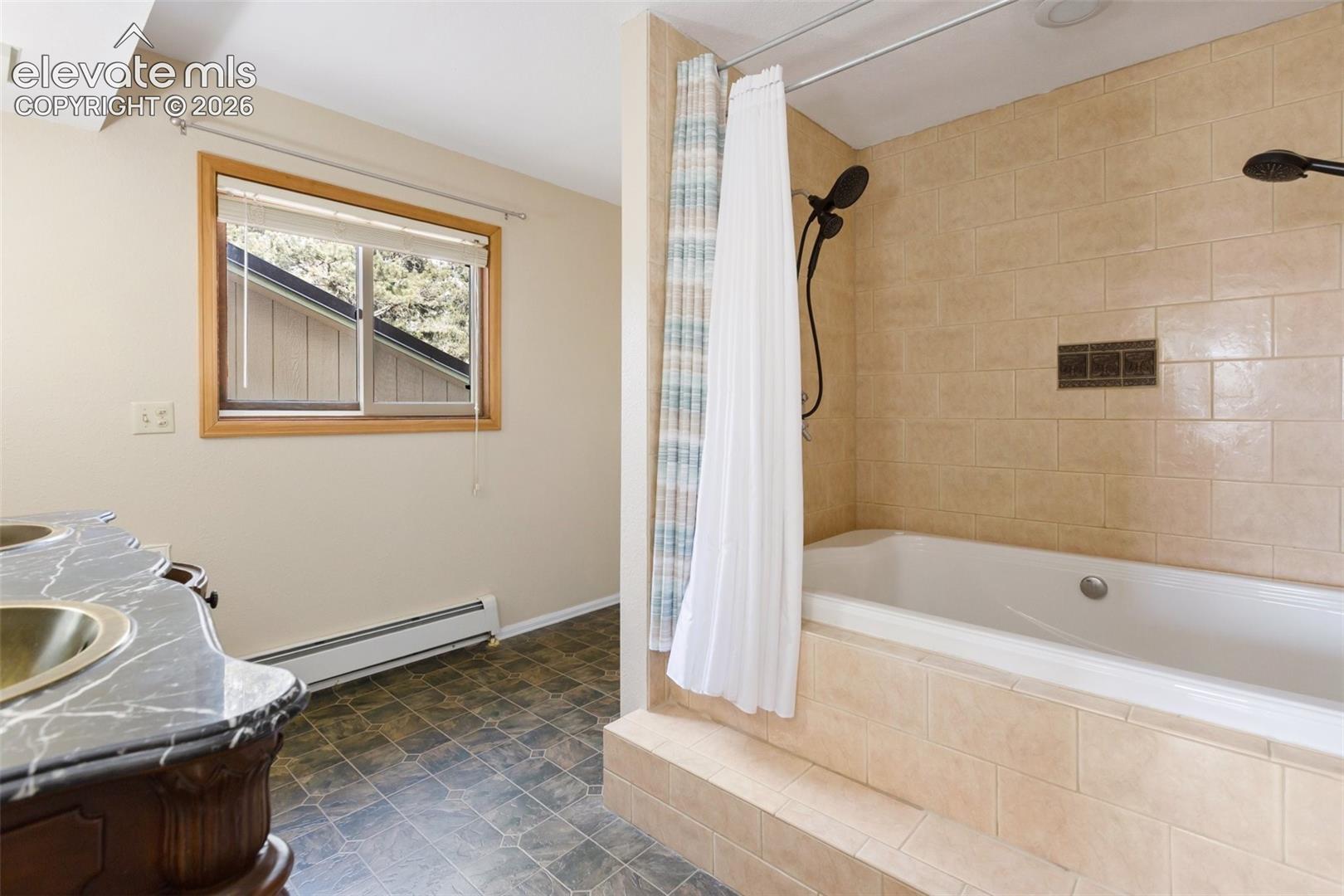 Tile-surround soaking tub with dual shower heads, wood-framed window, and a vanity featuring dual sinks with a dark countertop